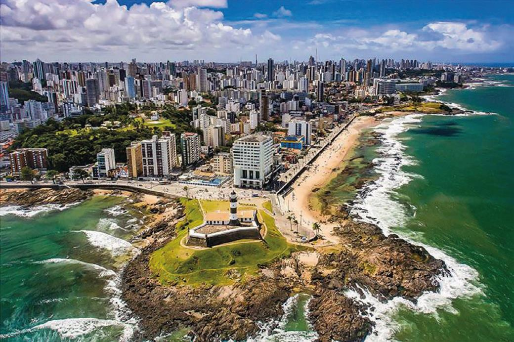 Fotografia aérea da orla da cidade de Salvador, capital da Bahia, que apresenta uma paisagem urbana composta pela presença de um farol, uma pequena área verde e muitos edifícios modernos, com diferentes alturas, localizados desde a área costeira até o interior da cidade.