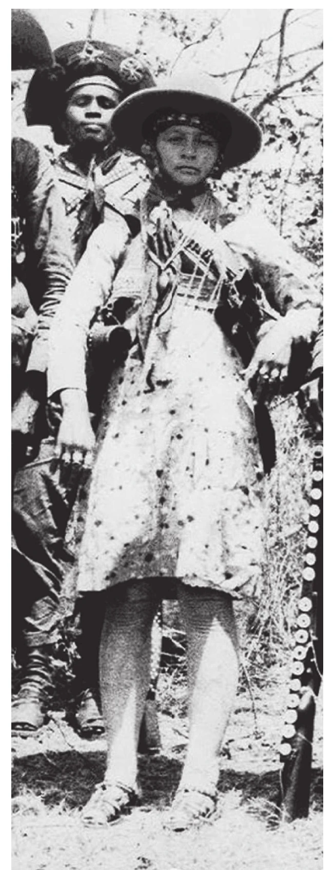 fotografia em preto e branco de Maria Bonita ao lado de um cangaceiro, de chapéu de couro adornado, vestido claro estampado, colares cruzados no peito e sandálias