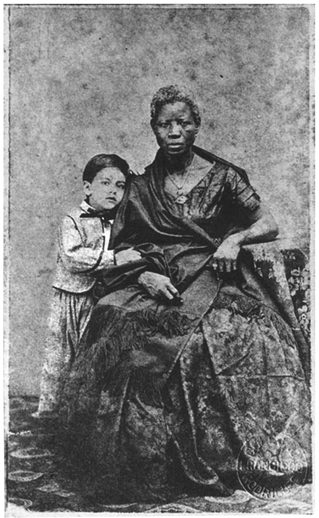 retrato de estúdio do século XIX: uma mulher negra sentada, de vestido longo com xale e colar, olha para a câmera; ao lado dela, em pé e encostado em seu braço, um menino branco de colete e gravata-borboleta
