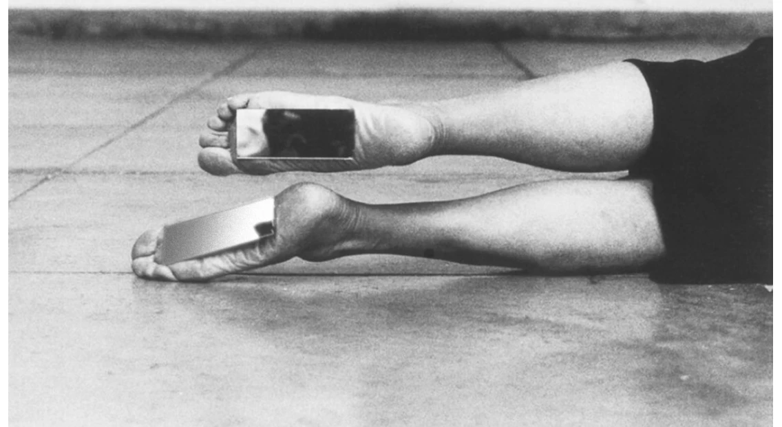 fotografia em preto e branco: as pernas e os pés descalços de uma pessoa deitada no chão de cimento, com pequenos espelhos retangulares apoiados sobre os pés, refletindo imagens escuras