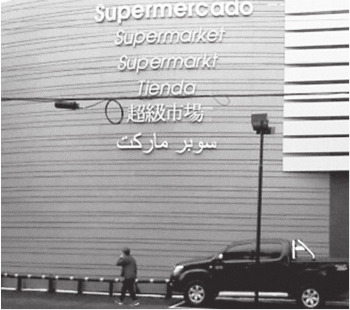fotografia da fachada de um supermercado em que o letreiro repete o nome do estabelecimento em seis idiomas, um abaixo do outro: “Supermercado”, “Supermarket”, “Supermarkt”, “Tienda”, em caracteres chineses e em árabe; embaixo, um pedestre caminha na calçada ao lado de uma caminhonete estacionada