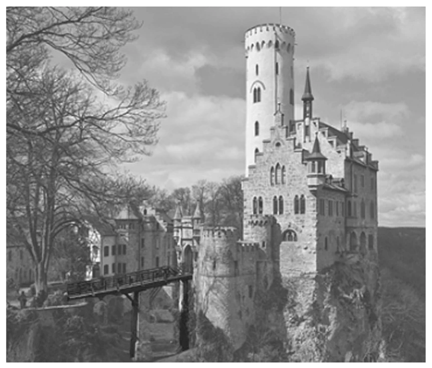 fotografia em preto e branco do castelo de Liechtenstein, erguido no alto de um paredão rochoso, com sua torre cilíndrica e telhados pontiagudos; uma ponte estreita de madeira sobre o abismo liga o castelo às construções vizinhas
